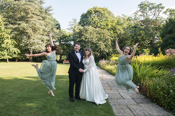 Bride and groom standing smiling in the center while bridesmaids jump joyfully around them in the Botanical Gardens, Bath